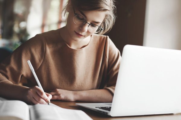 A focused woman in glasses writing notes while researching legal documents on a laptop.