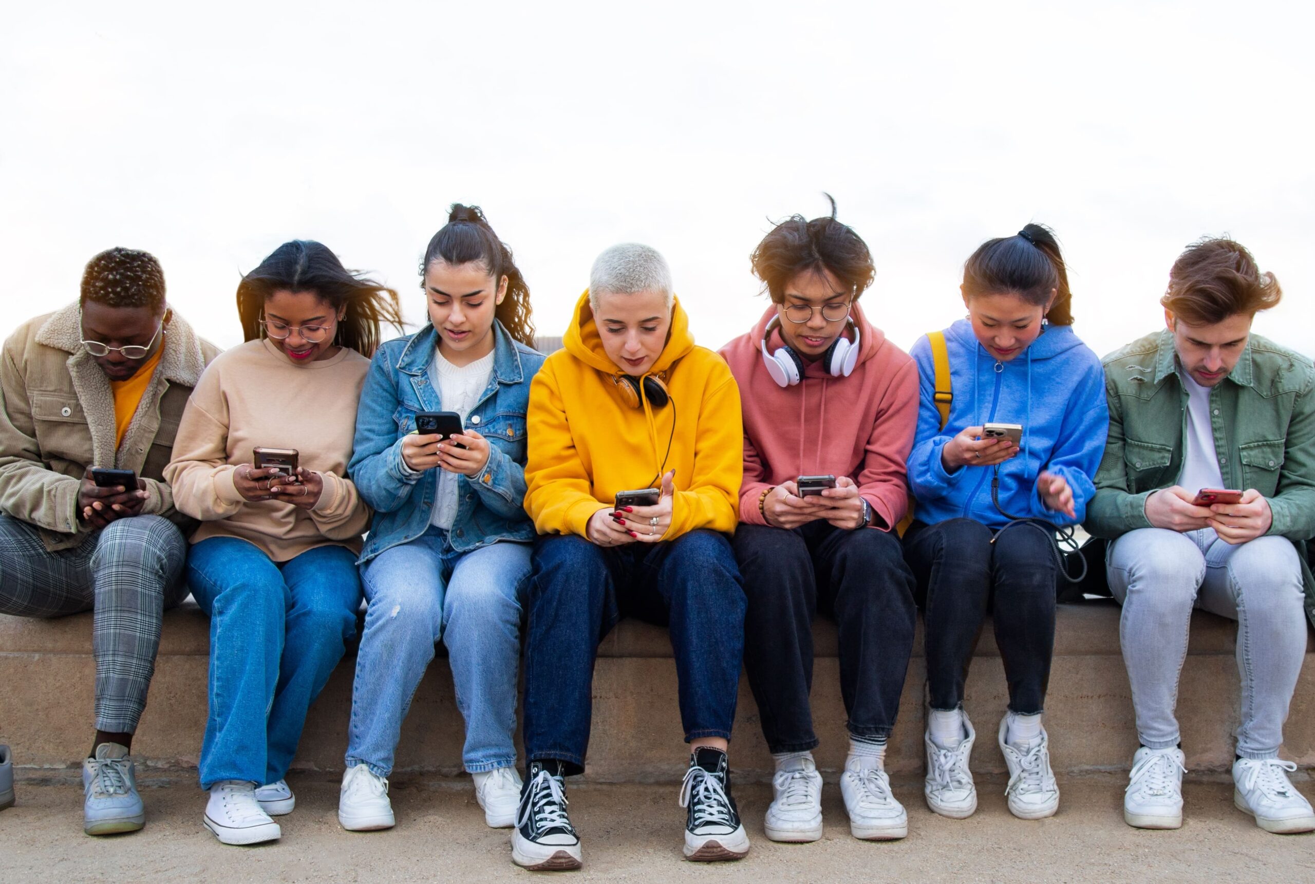 Group of teenagers and young adults sitting on a bench, engrossed in their smartphones. Lawsuits claim social media platforms contribute to addiction and mental health issues.