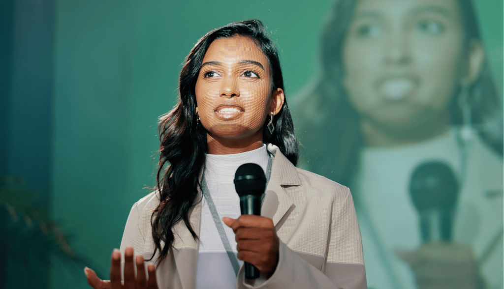 A confident woman in a beige blazer speaking into a microphone, sharing her success story.
