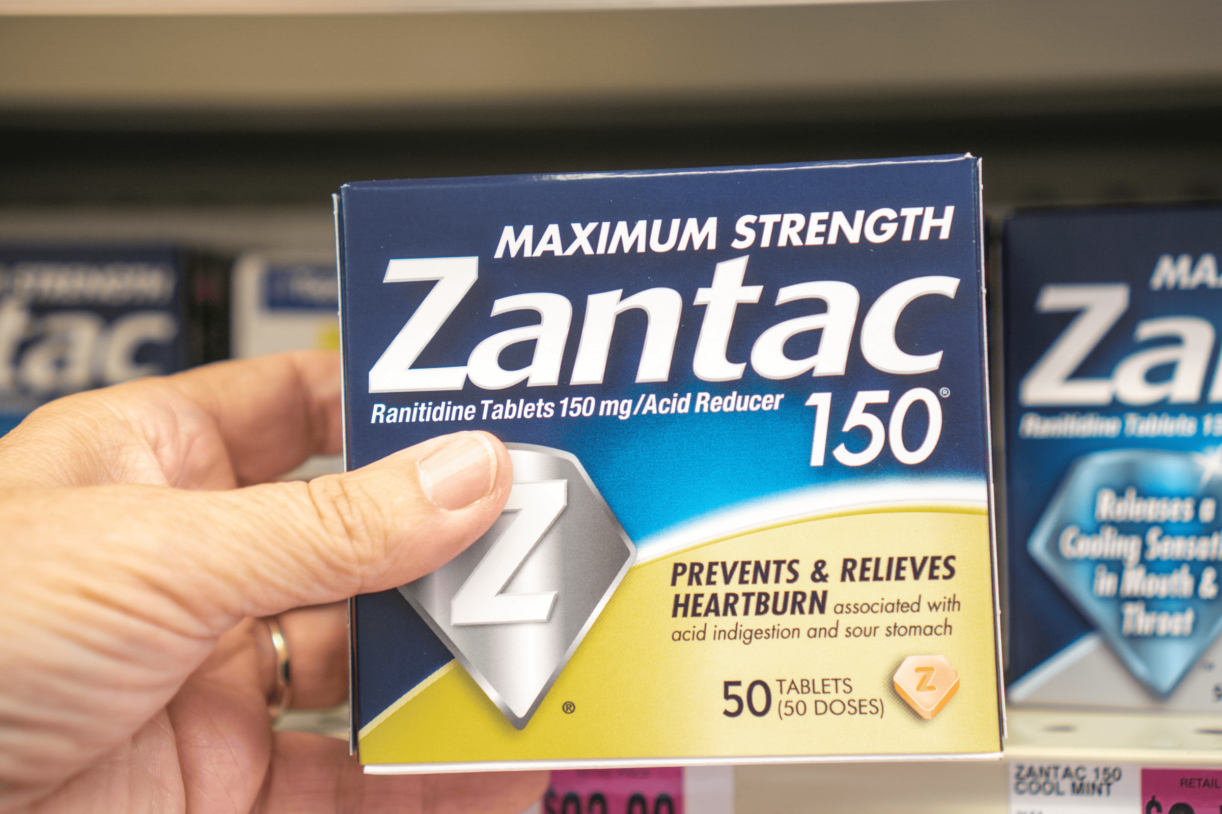 A hand holding a box of Zantac 150 heartburn medication on a store shelf, linked to lawsuits over cancer risks.
